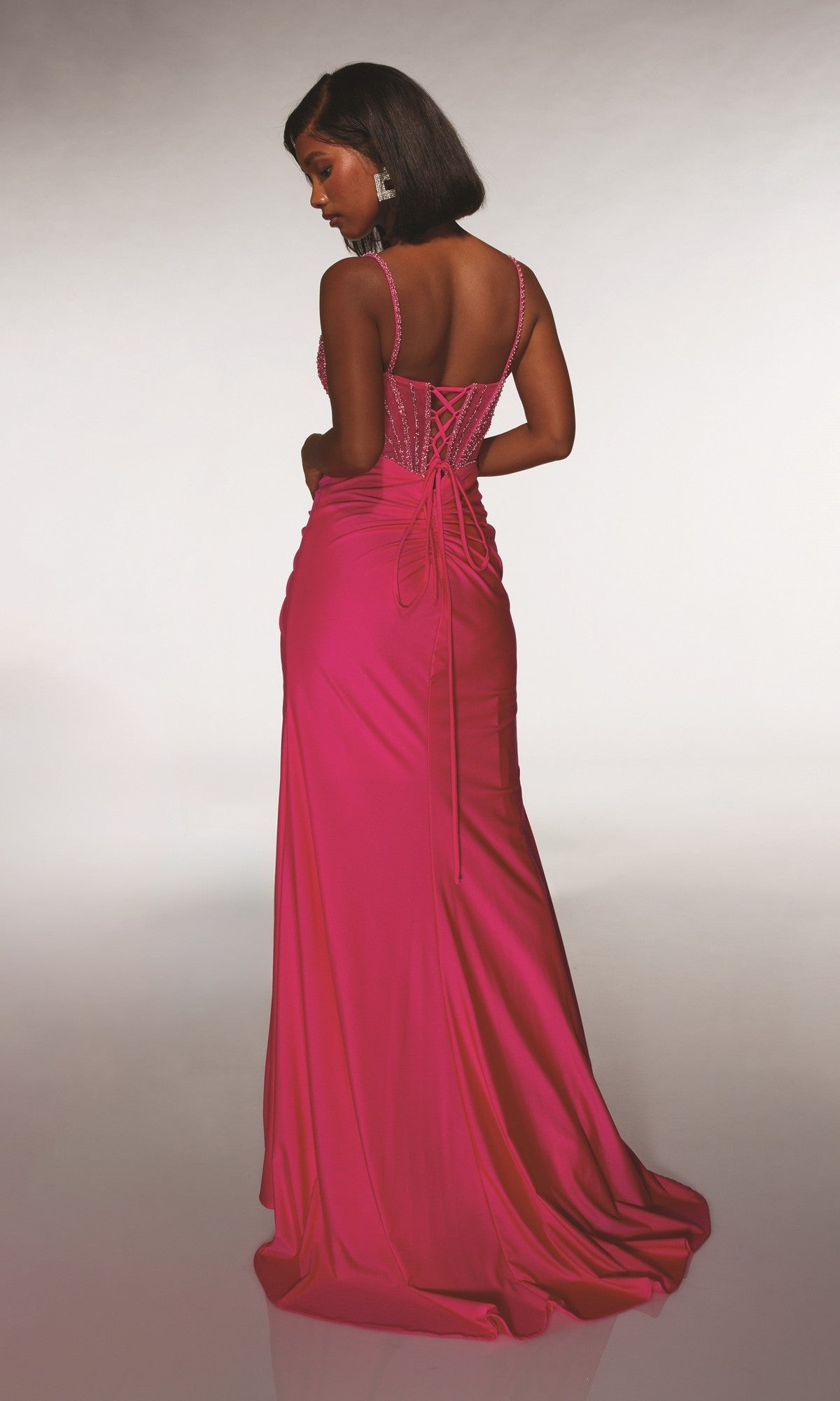 A woman stands with her back to the camera, wearing the Alyce 62113 long prom dress in bright pink. The floor-length, sleeveless gown features a beaded corset bodice and a lace-up open back.